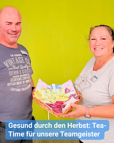 Eine Kollegin überreicht ein Körbchen mit Tee an einen Kollegen. Beide lächeln in die Kamera. Der Hintergrund ist leuchtend grün. Bildbeschriftung: „Gesund durch den Herbst: Tea-Time für unsere Teamgeister“